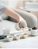 Person pouring tea from a decorative teapot into small cups on a light surface.