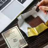 Fossa Chocolate Nanyang Kopi with a hand holding a piece of chocolate and a laptop in the background.