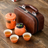Tea set with orange teapot and cups on a wooden surface next to a brown leather suitcase shaped cover.