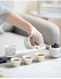 Person pouring tea from a decorative teapot into small cups on a light surface.