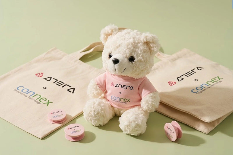 A cream-colored plush teddy bear wearing a pink shirt with the 'Ategra + Connex' logo sits between two matching beige tote bags on a light green background. Small pink heart-shaped accessories are scattered around the products.