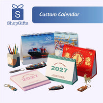 A promotional display of custom 2027 desk calendar products and matching keychains on a light lavender background. The collection includes various designs: a red Chinese New Year calendar with gold lion illustrations, a green and white New Year calendar, a blue cargo ship calendar, and other pink and multicolor options.
