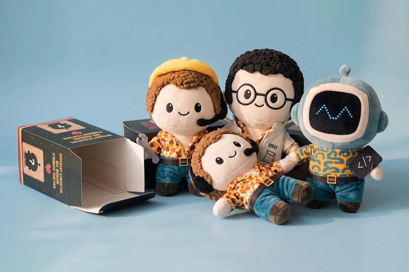 Three plush toy figures and an open product box on a light blue studio background. The toys have cartoon-style faces, one with brown hair and a yellow cap, one with dark hair and glasses, and one with a blue robot head.