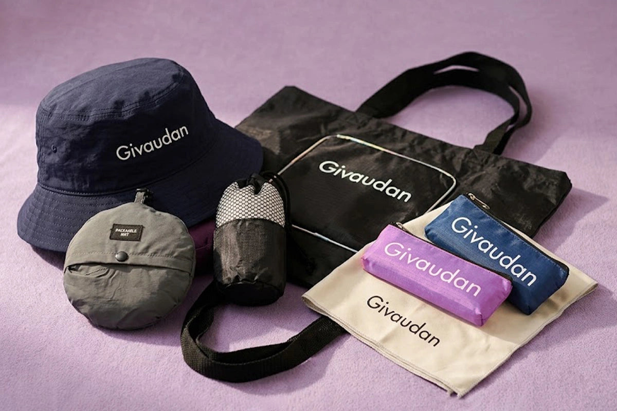 A Givaudan-branded camping gear set arranged on a lavender background, including a navy bucket hat, a black tote bag, a gray compact pouch, a black-and-white mesh cup holder, a purple pouch, and a blue pouch on a cream drawstring bag.