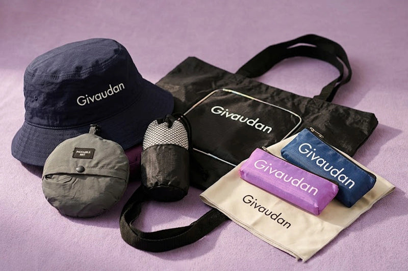 A Givaudan-branded camping gear set arranged on a lavender background, including a navy bucket hat, a black tote bag, a gray compact pouch, a black-and-white mesh cup holder, a purple pouch, and a blue pouch on a cream drawstring bag.
