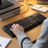 An employee typing on a black Logitech K120 USB keyboard at a corporate desk cubicle, a dependable tech asset for new hire onboarding kits. Customised Corporate Gift