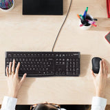 A remote professional using the full-size Logitech K120 wired keyboard on a wooden home office desk with an external monitor setup_Custom Keyboard Corporate gift