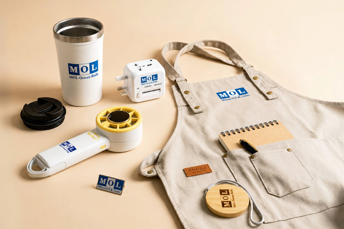 A styled flat-lay of MOL-branded office accessories on a warm beige background, including a beige apron with pockets, a white insulated travel mug, a white power adapter, a white handheld device with yellow accents, a small notebook, a pen, and a wooden round accessory.