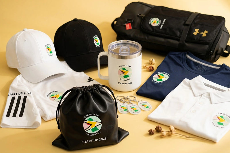 A coordinated merchandise set arranged on a warm beige background, including a black drawstring pouch, a black cap, a white cap, a white t-shirt with black stripes, a white t-shirt with a circular logo, a white polo shirt, a navy polo shirt, a white tumbler, and a black zip pouch. The items feature a South Africa-themed circular logo and text reading 'Start Up 2025' and 'Start Up 2025 TE Connectivity'.