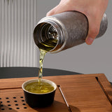 Hand pouring liquid from a metal container into a small cup on a wooden surface.