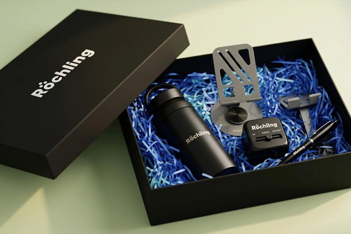 Röchling gift set in an open black box with blue paper shred, featuring a black insulated bottle, metal utensils, a small black accessory, and a pen on a pale green background.