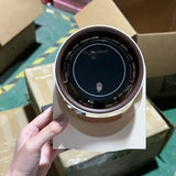 Hand holding a cylindrical electronic device with a dark circular component on a cardboard surface.