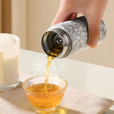 Hand pouring tea from a Titanium bottle into a glass cup on a neutral background