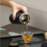 Person pouring tea from a black and silver tea infuser into a glass cup on a textured surface.