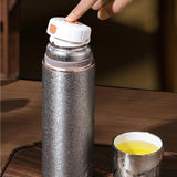 Titanium Thermal Brew Mug with white cap on a wooden surface, next to a cup of tea.
