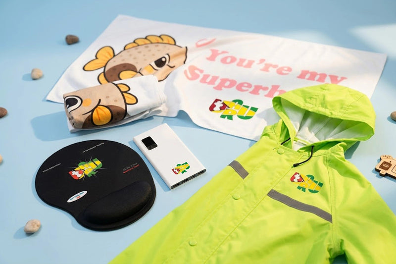 A flat-lay product display featuring a neon yellow-green hooded raincoat, a white towel with a cartoon fish and pink text, a matching white pouch, a black oval patch, and a white rectangular case, all decorated with a colorful frog logo, arranged on a light blue background.