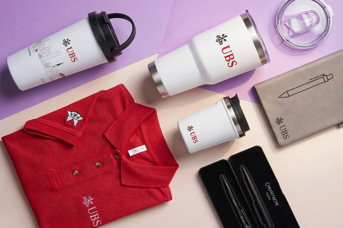 Collection of branded items including a red polo shirt, white tumblers, and a pen on a light purple background.
