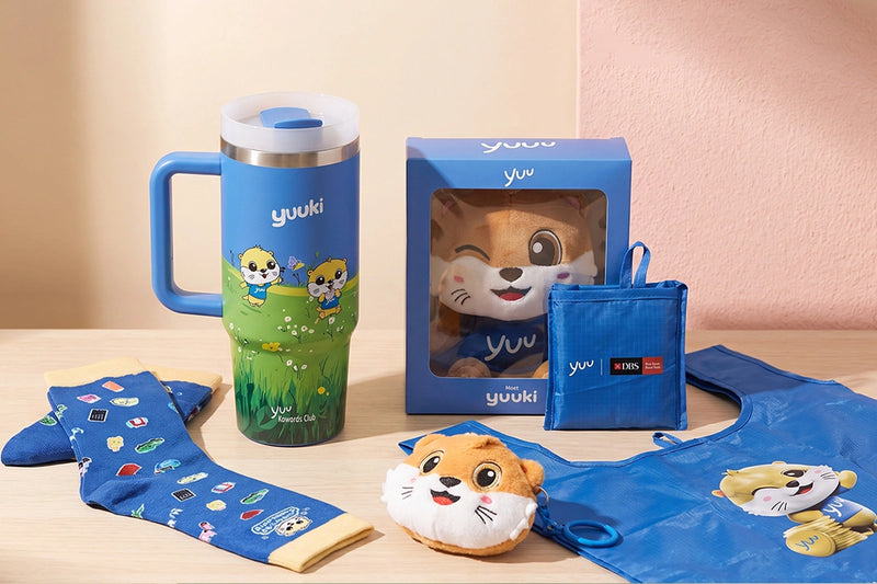 A Yuuki-branded gift set arranged on a tabletop against a soft beige and peach background. The set includes a blue handled tumbler with a green illustrated lower section, a blue plush toy in a clear window box, a blue zip pouch, patterned socks, a blue bib, and a small plush character.