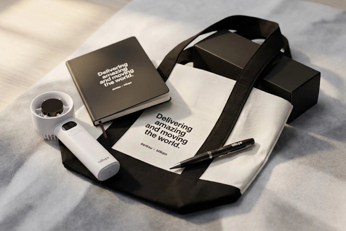 A styled flat-lay of a white tote bag with black handles and black printed text, alongside a black notebook, a white handheld device, a black pen, and a black box, all arranged on a light gray fabric surface.