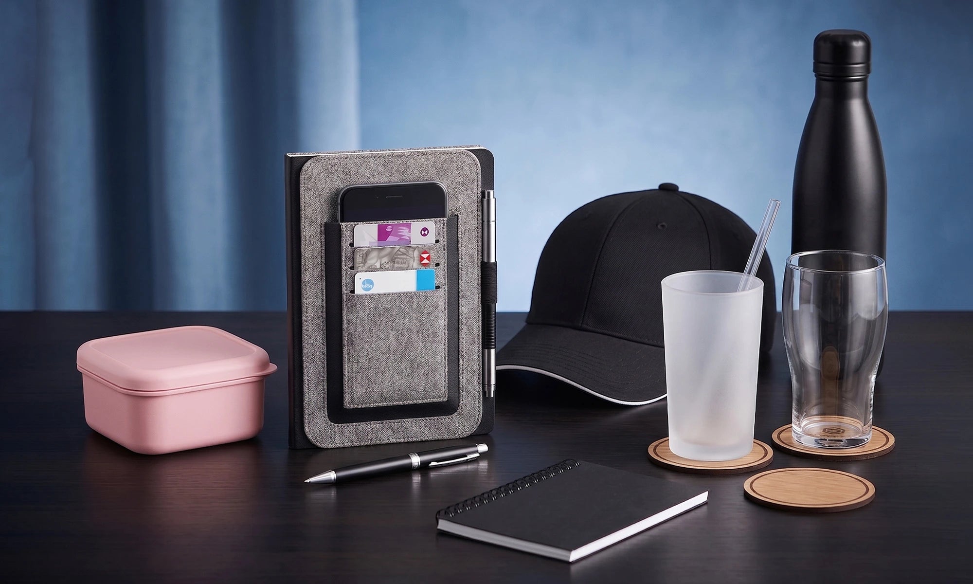 Collection of office supplies including a phone case, cap, notebook, and drinks on a dark surface with a blue gradient background.