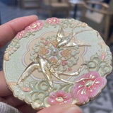 Decorative mirror with floral and bird design held by a hand.